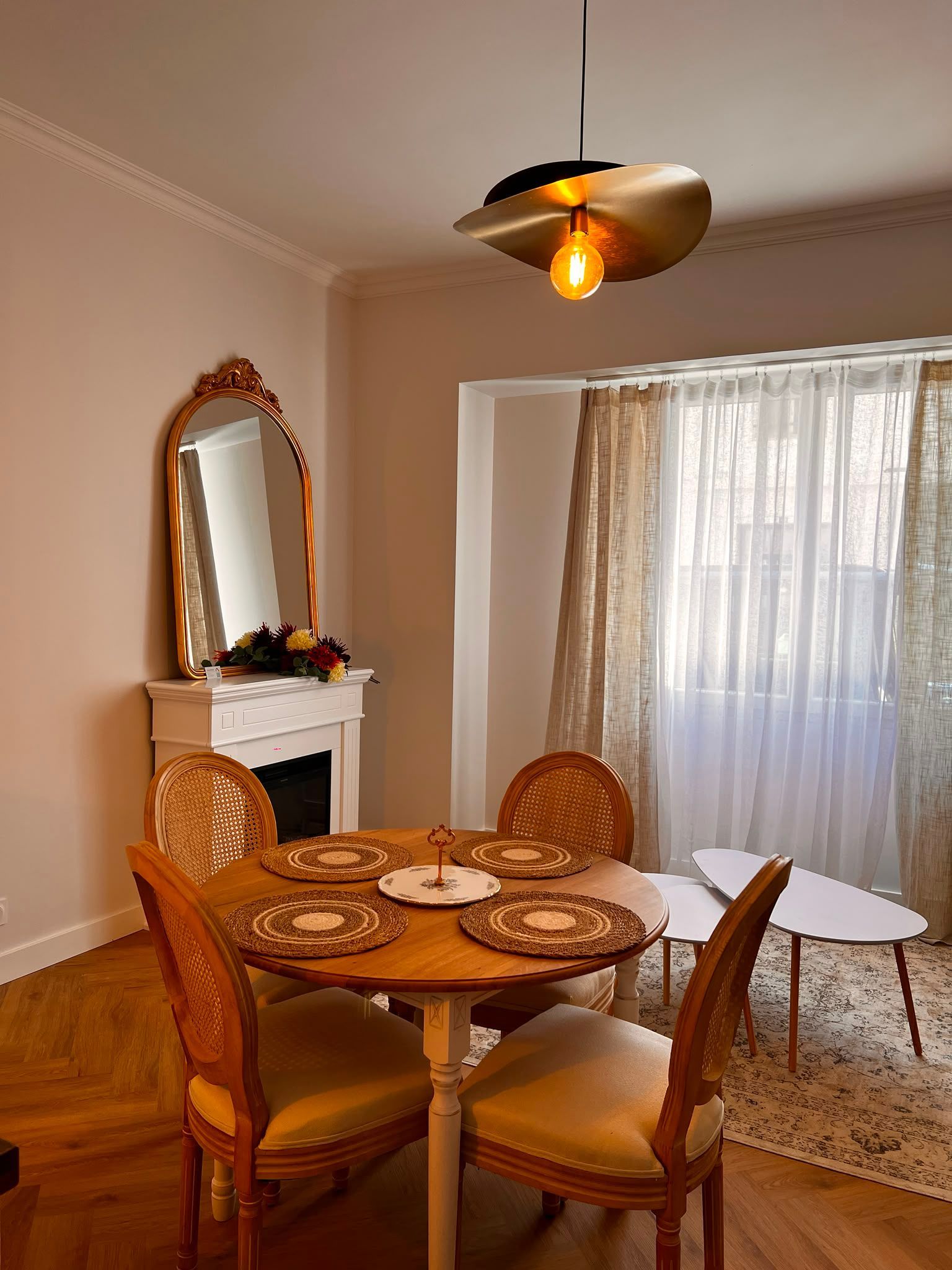Salle à manger avec table ronde en bois, quatre chaises, miroir, cheminée et grande fenêtre à rideaux.