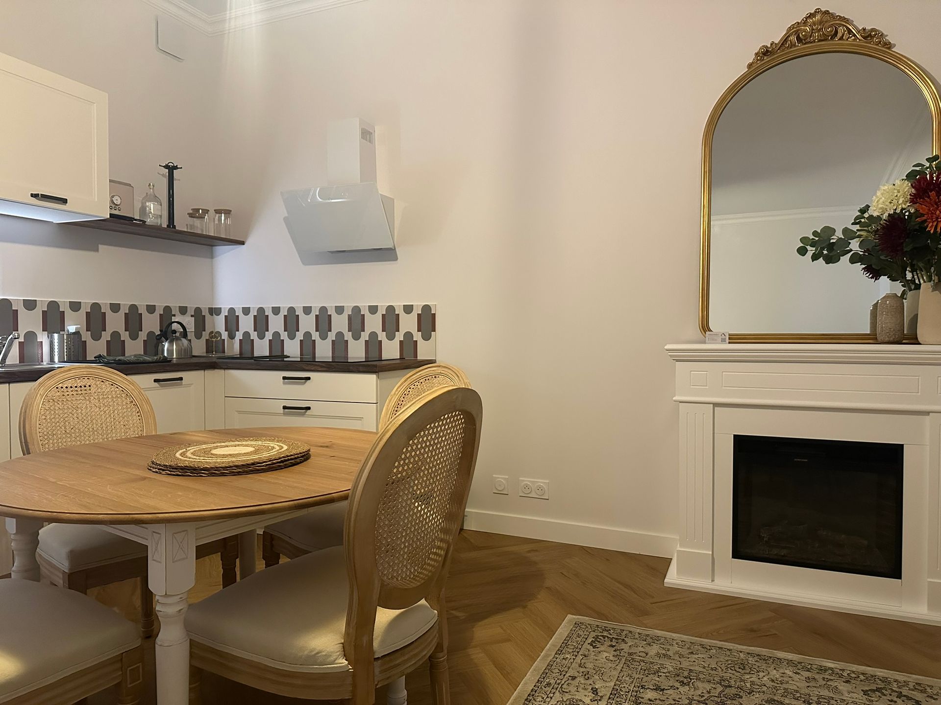 Salle à manger élégante avec table ronde en bois, chaises en osier et cheminée blanche ornée d'un miroir à cadre doré
