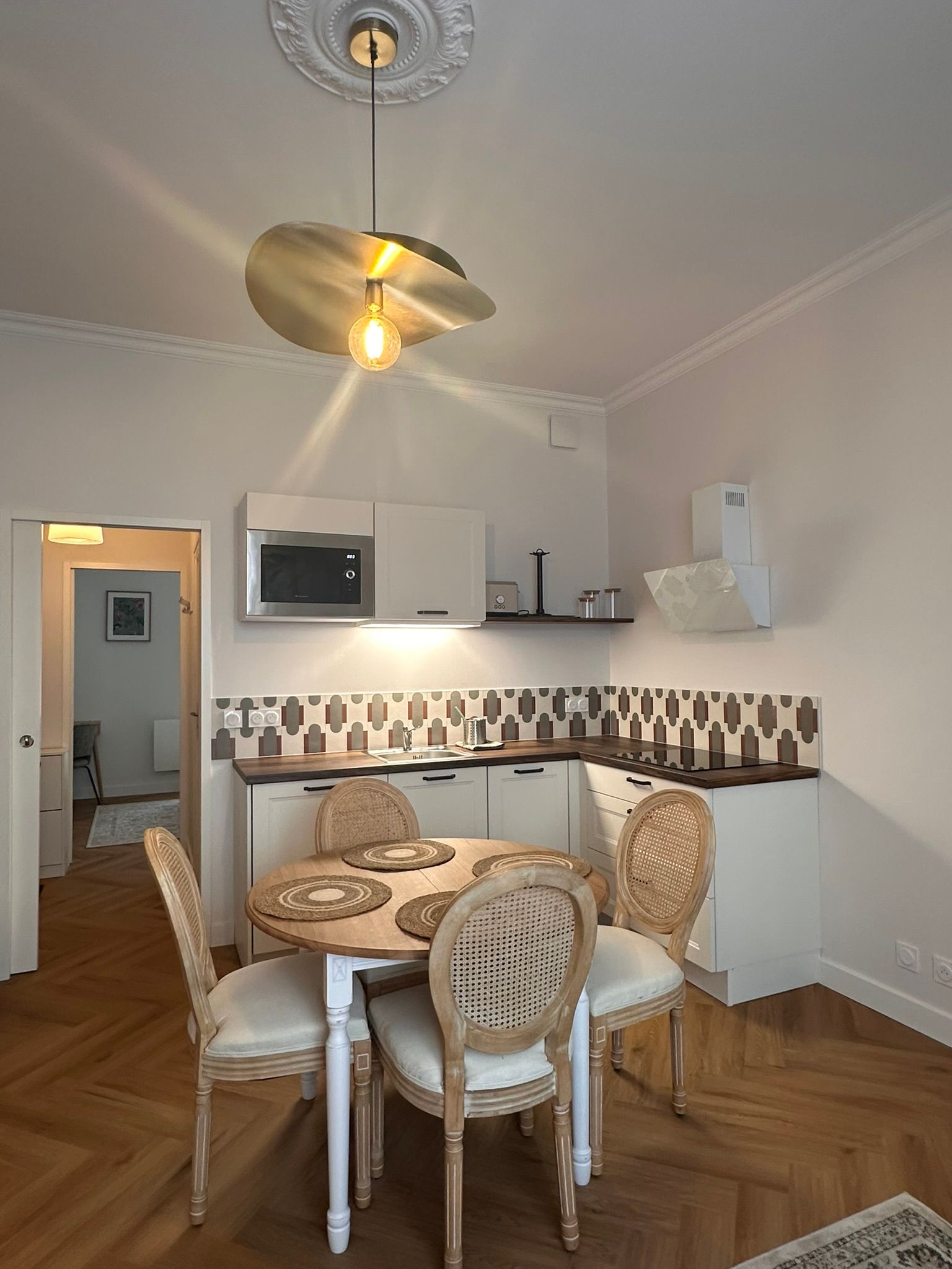 Petite cuisine lumineuse avec table ronde, chaises en osier, crédence à motifs et ventilateur de plafond.