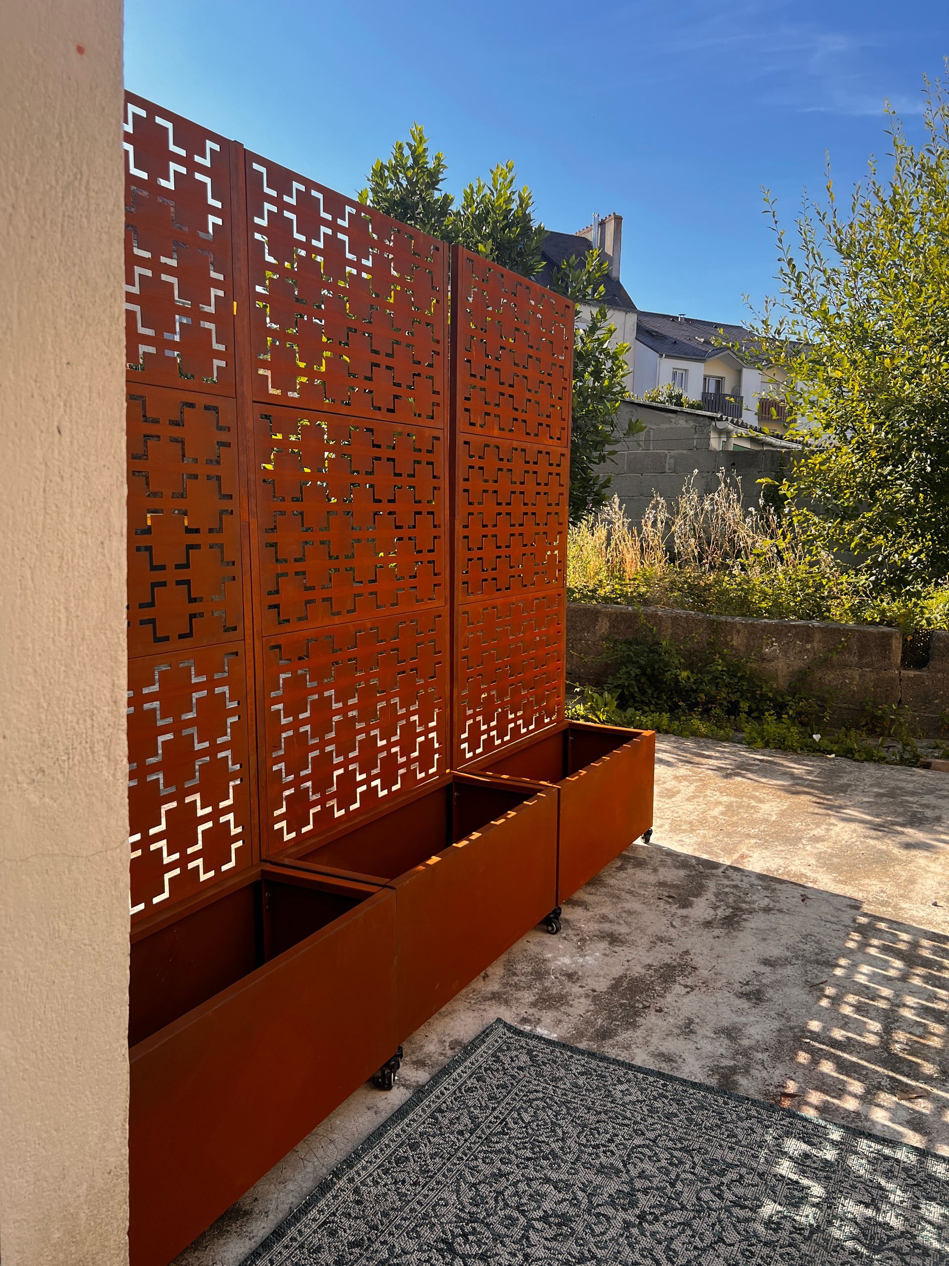 Panneau d'intimité en métal perforé couleur rouille, bordant un chemin de jardin en plein soleil.
