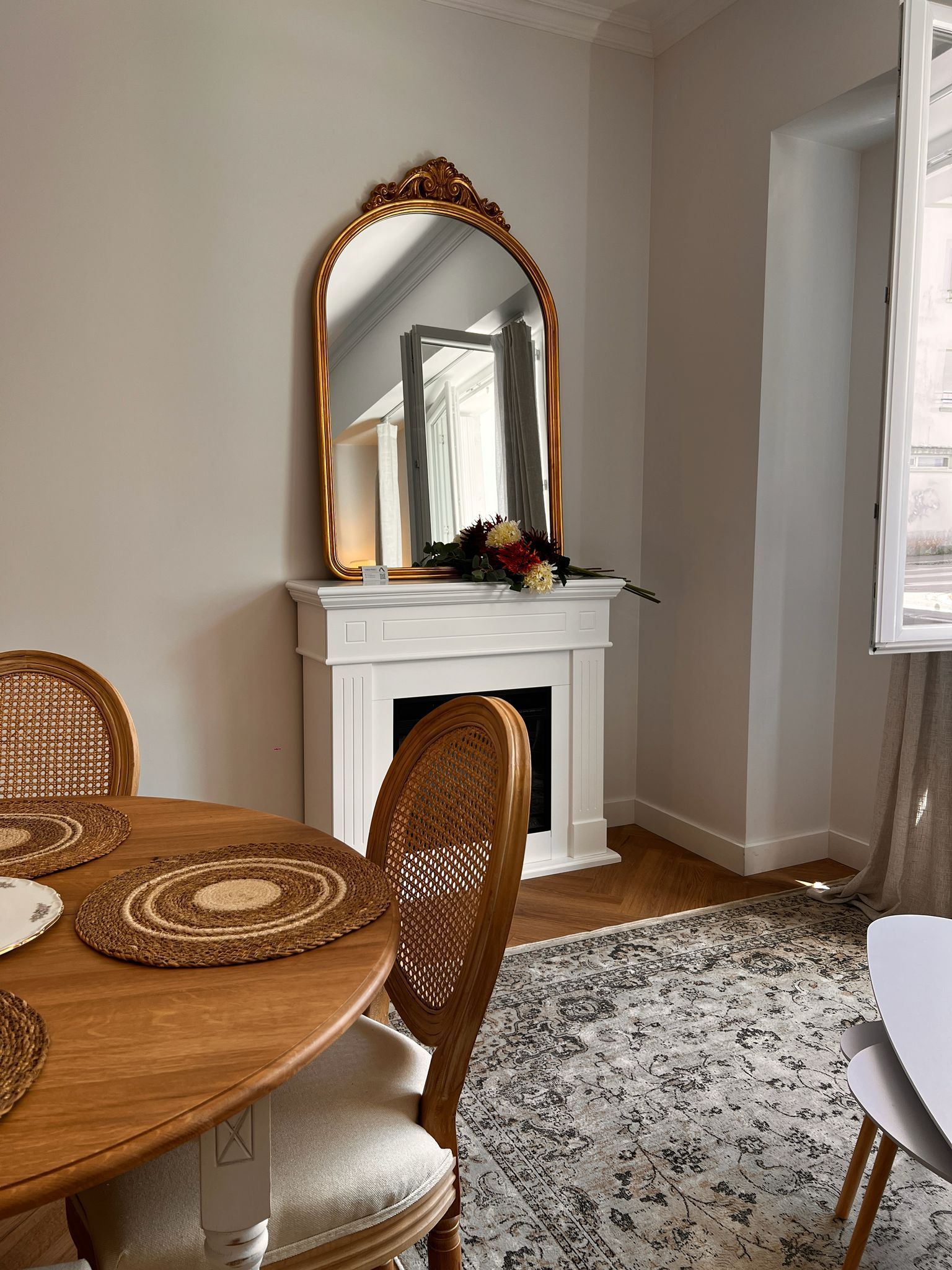 Coin repas avec une table ronde en bois, des chaises en osier, un miroir orné sur une cheminée blanche et un tapis à motifs