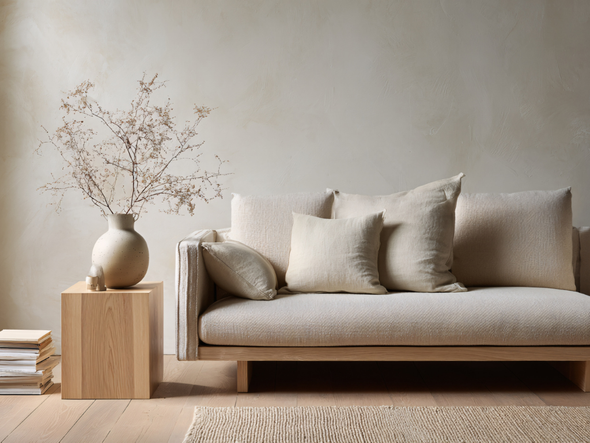 Canapé beige avec coussins, table d'appoint en bois, vase avec branches et mur neutre.