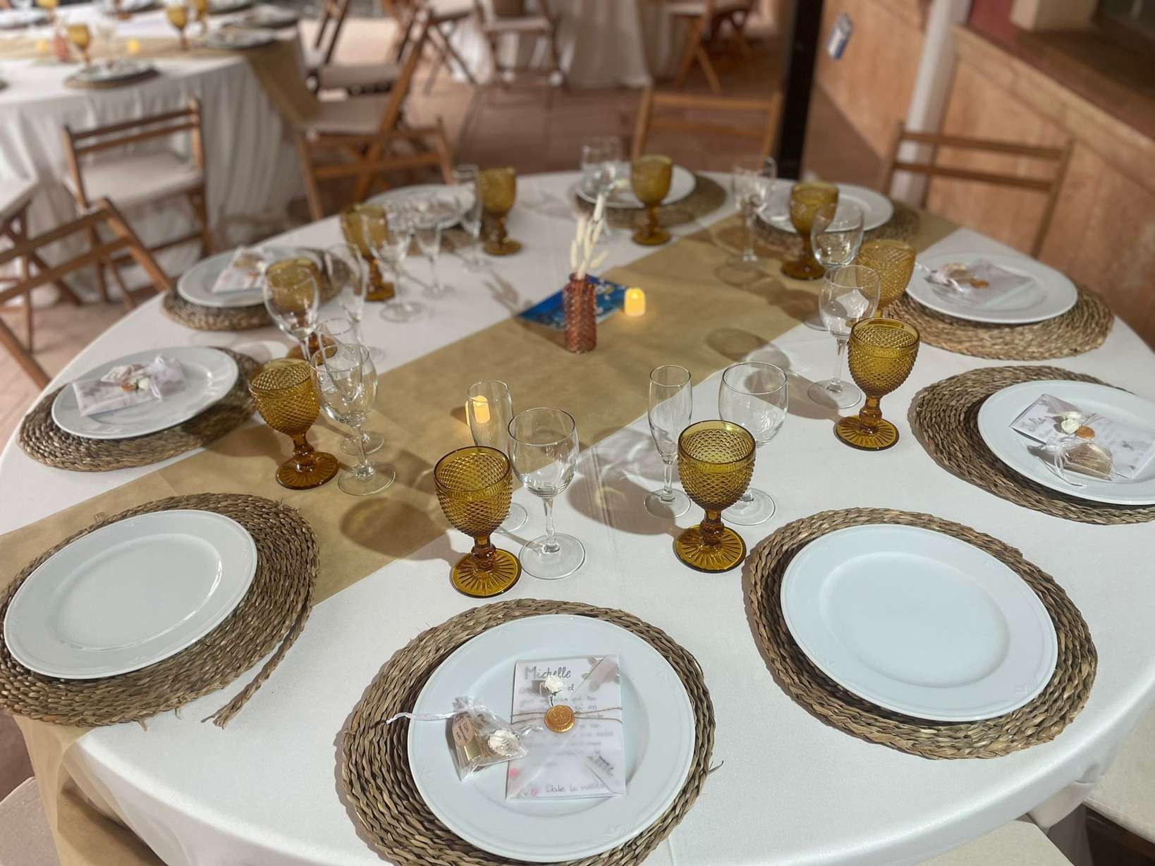 Mesa redonda para evento formal con mantel blanco, manteles individuales tejidos y cristalería color ámbar.