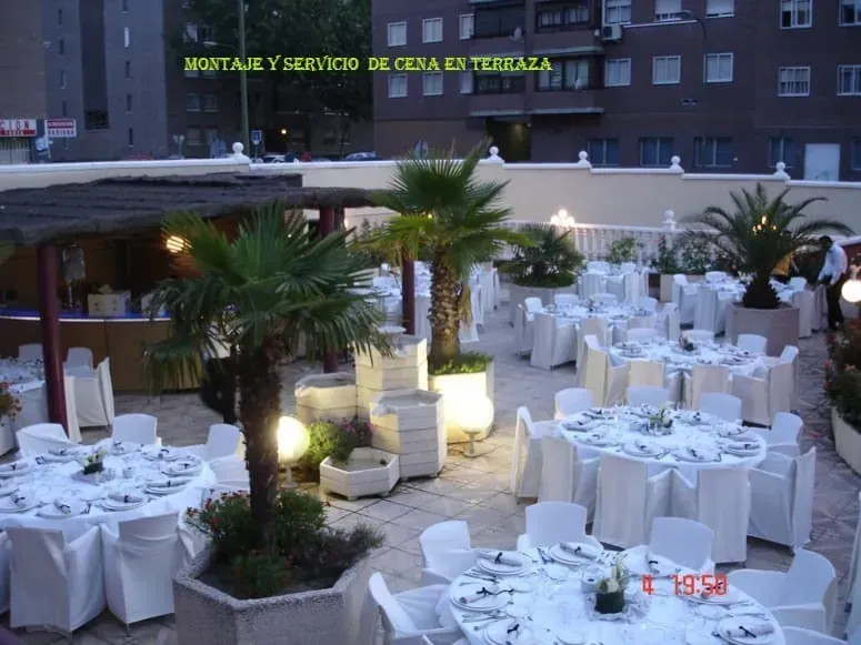 Cena al aire libre con mesas y sillas cubiertas de blanco y palmeras, iluminadas con una luz suave.