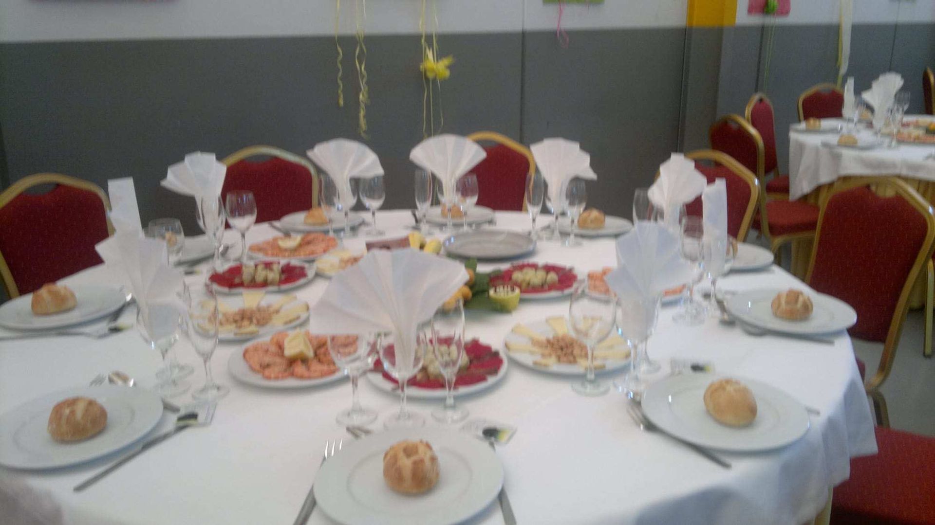 Mesa redonda dispuesta para comer, con platos, comida, vasos y sillas con respaldo rojo en una habitación.