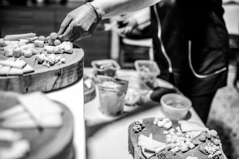 Persona colocando comida en una mesa, incluyendo queso y uvas. Blanco y negro.