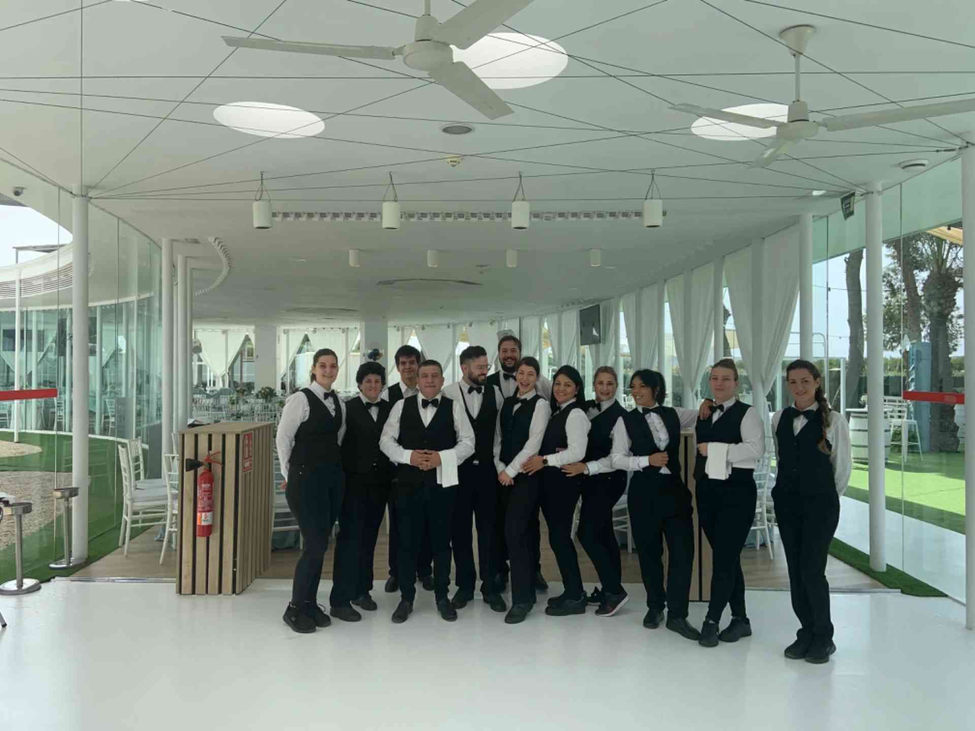 Grupo de camareros con chalecos negros y pajaritas, posando dentro de un restaurante moderno y luminoso.
