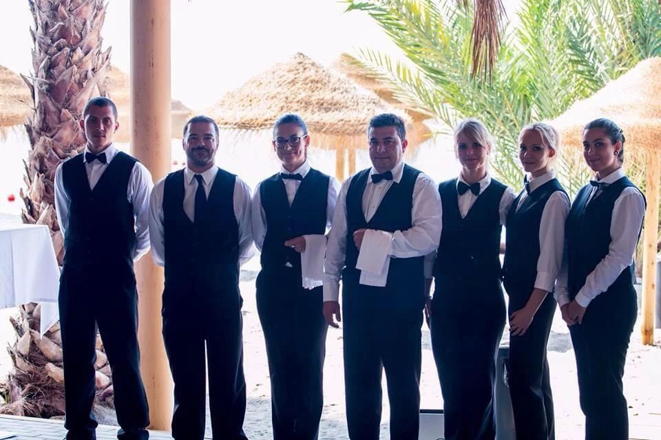 Grupo de siete camareros con chalecos negros, camisas blancas, pajaritas y pantalones negros, al aire libre.