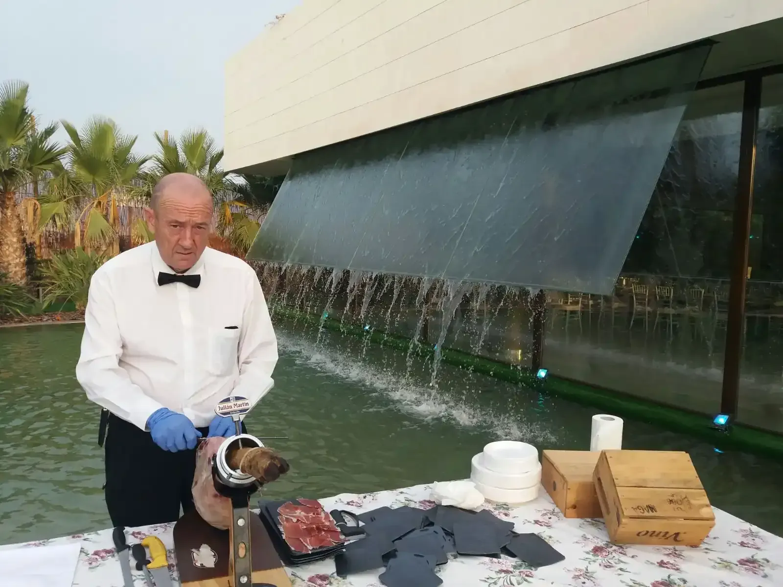 Hombre cortando carne, con pajarita, junto a una cascada y una fuente de agua.