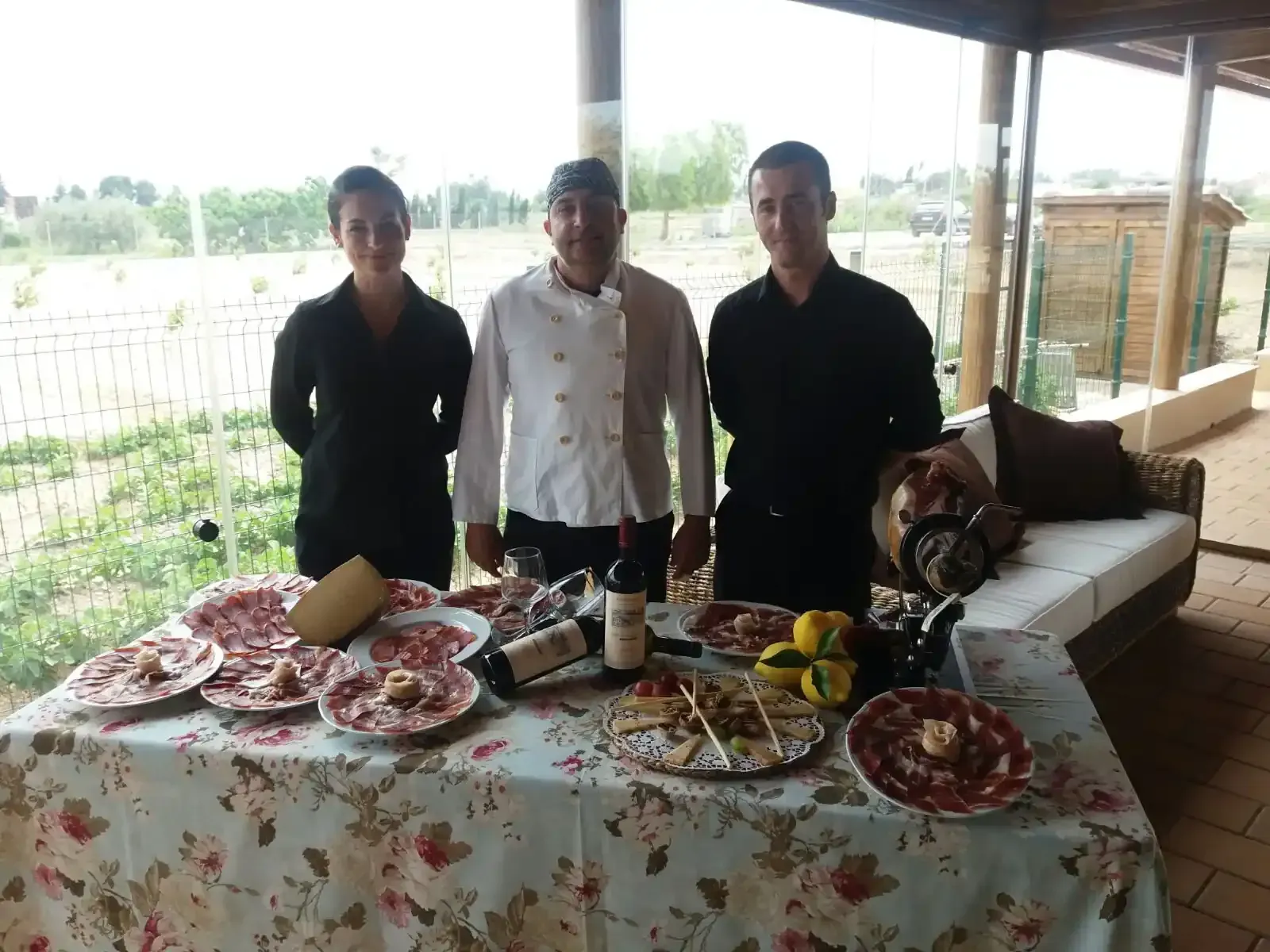 Tres empleados del restaurante posan tras una mesa al aire libre. Se exhiben fiambres, vino y queso.