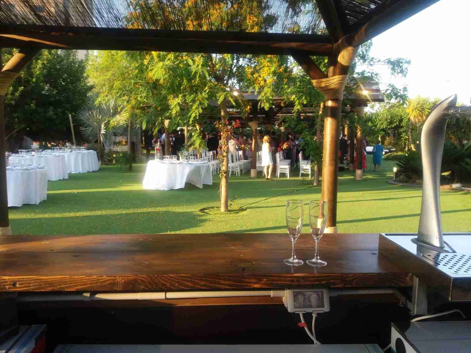 Bar con copas de champán con vistas a un césped con mesas de boda e invitados bajo un cenador de madera.