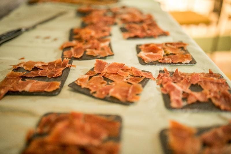 Rebanadas de carne curada dispuestas en pequeños platos cuadrados de color negro, sobre una superficie blanca.