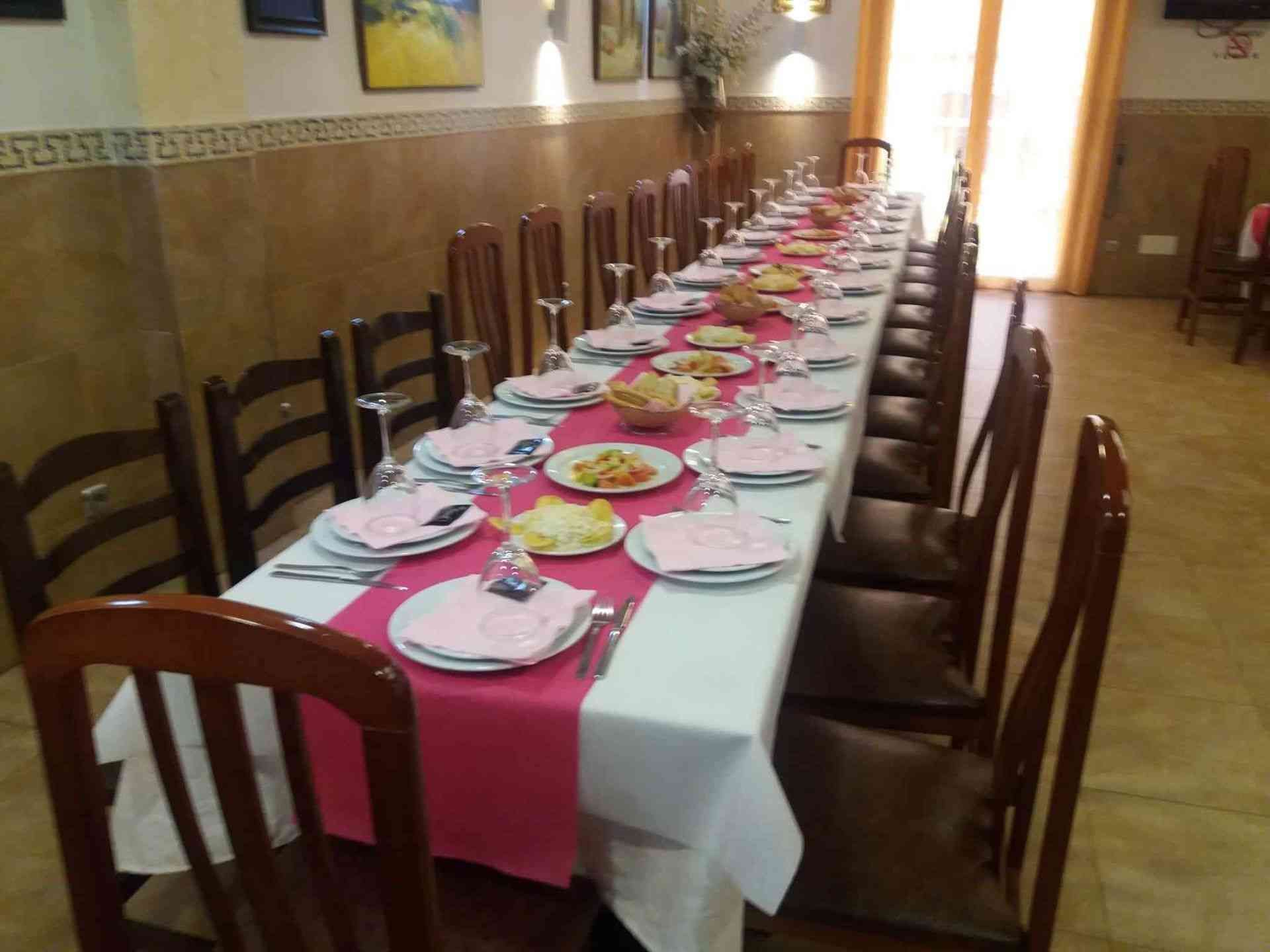 Mesa de comedor larga para comer, con mantel blanco, camino de mesa rosa y cubiertos.