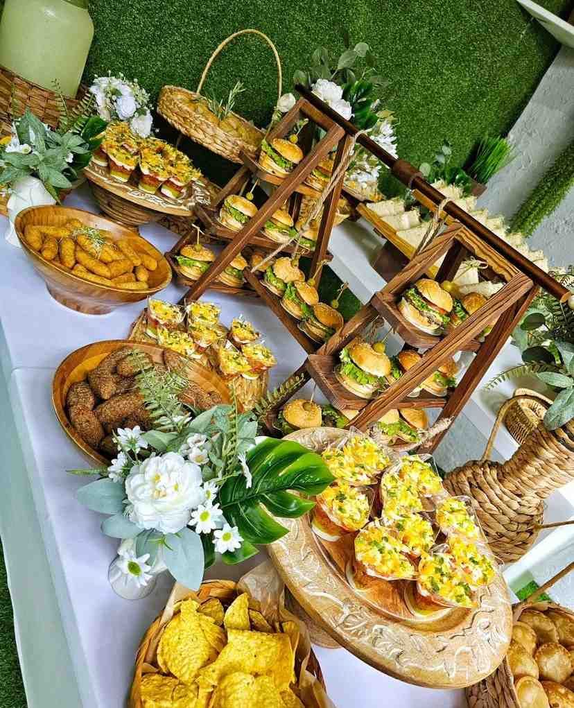 Mesa de comida con snacks: mini hamburguesas, patatas fritas, buñuelos y bollería, sobre mantel blanco, decorada con flores.