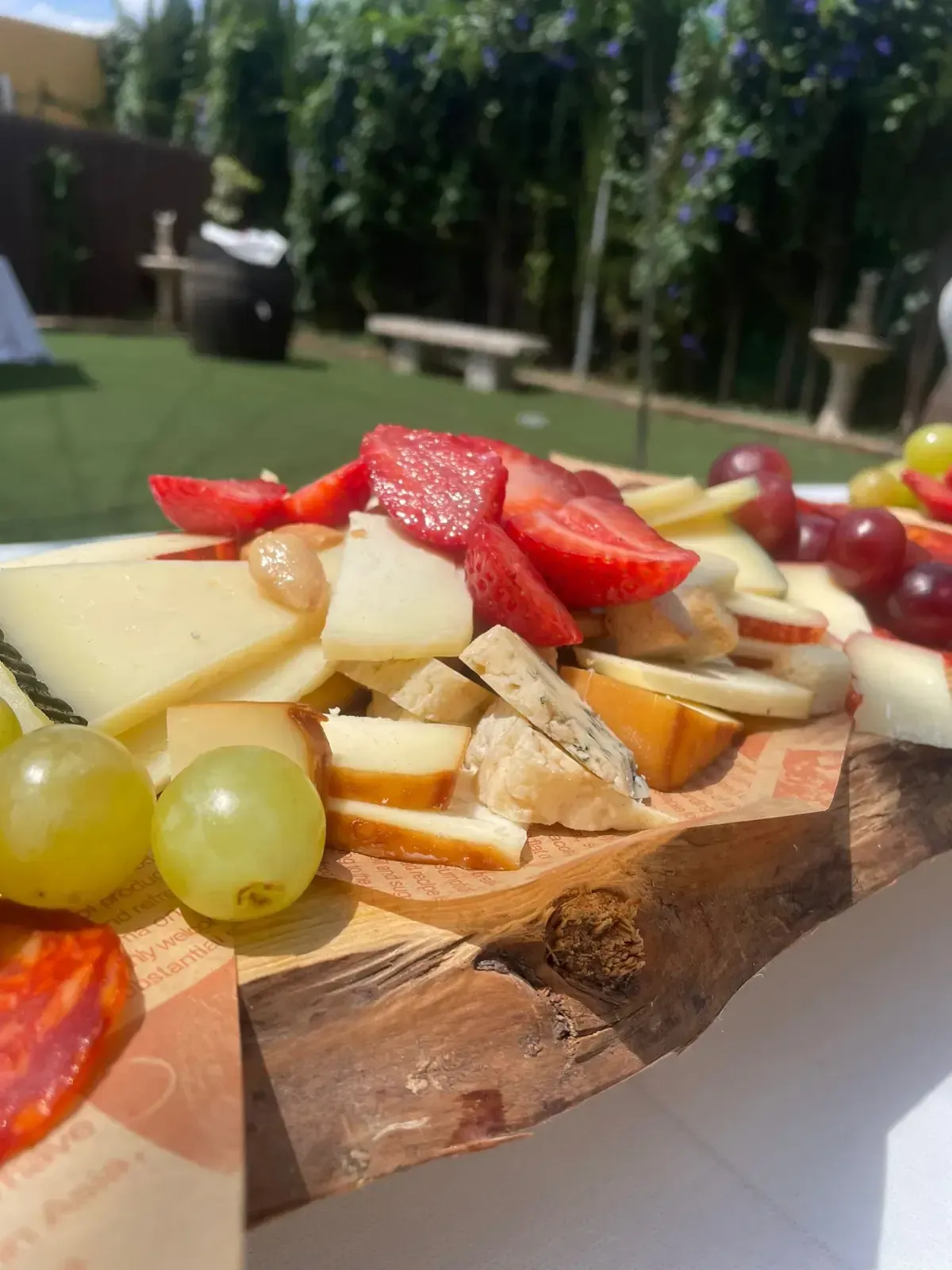 Tabla de embutidos con queso, fresas, uvas y rodajas de manzana, al aire libre en un día soleado.