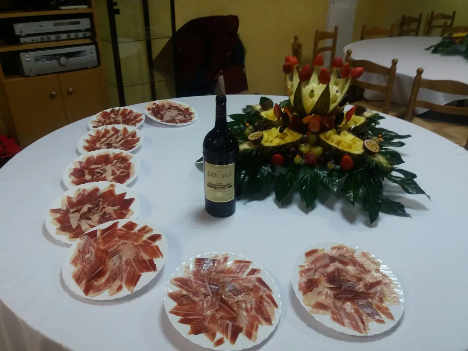 Mesa puesta con platos de jamón lonchas, botella de vino y centro de fruta.