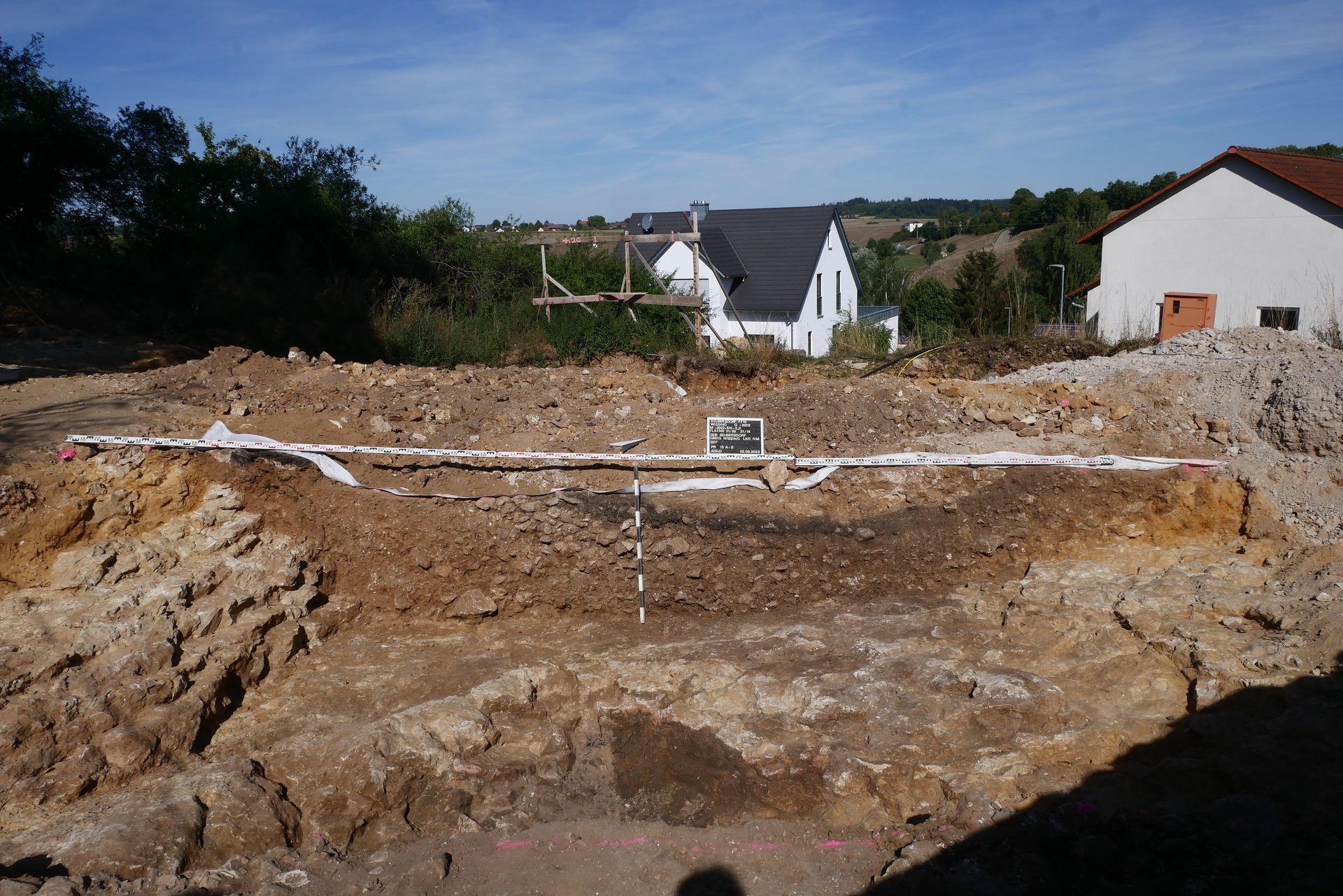 Ausgegrabene archäologische Stätte mit geschichteter Erde, Messstäben und Gebäuden im Hintergrund.