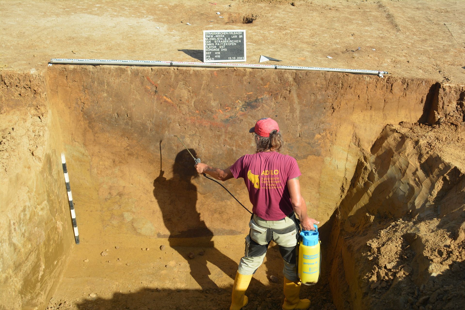 Archäologe sprüht Wasser auf ein Bodenprofil in einer Baugrube; braune, hellbraune und rote Erde.