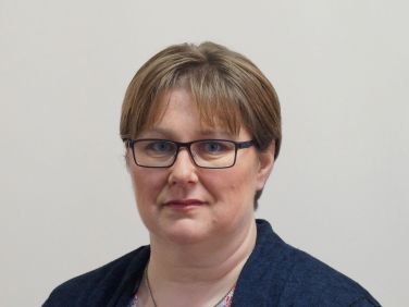 Frau mit Brille und kurzen braunen Haaren, trägt eine dunkelblaue Jacke und blickt in die Kamera.