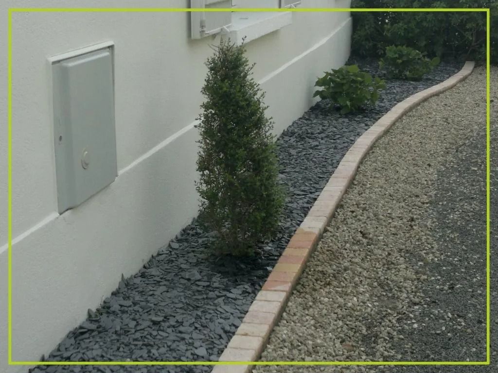 Un lit de gravier foncé et de petites plantes jouxte un bâtiment de couleur claire, bordé d'un mur de briques et d'un chemin de gravier