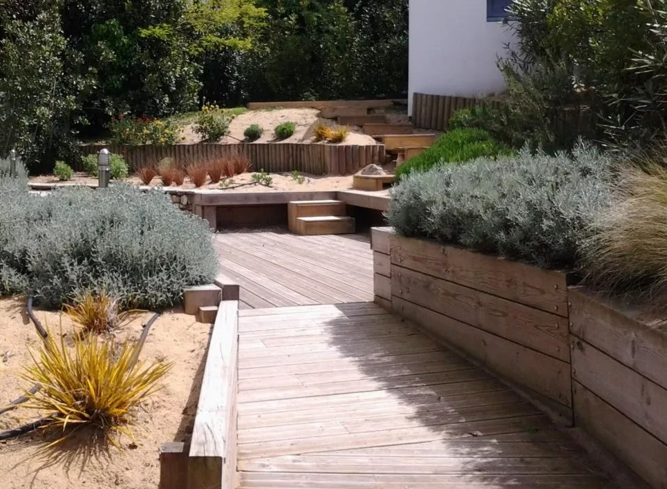 Un chemin en bois mène à une cour paysagée avec des bancs intégrés, agrémentée de diverses plantes et de murs de soutènement