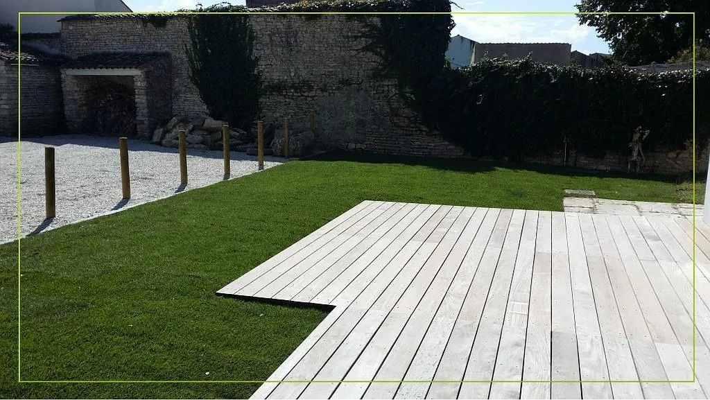Terrasse en bois jouxtant une pelouse verdoyante, mur de pierre avec végétation en arrière-plan