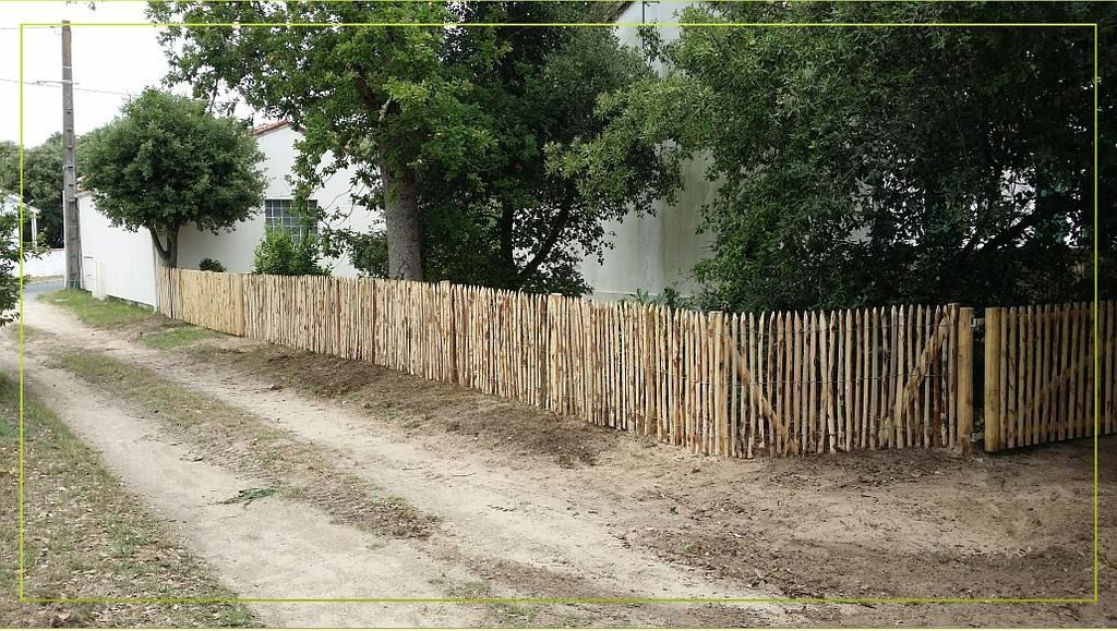 Une clôture en bois borde un chemin de terre, des arbres et une maison