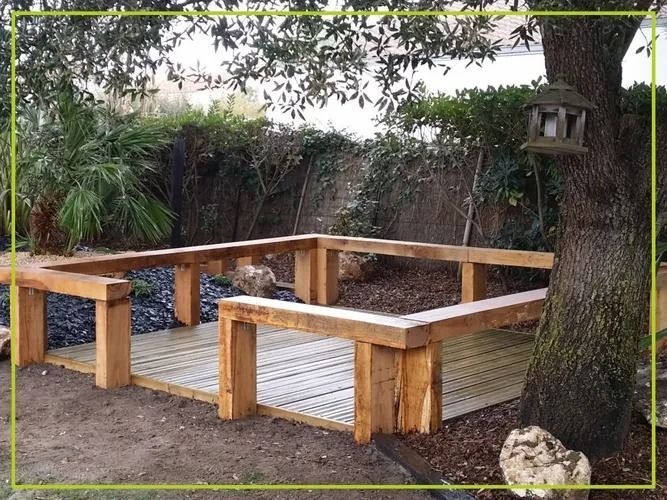 Terrasse en bois avec bancs intégrés sous un arbre, entourée d'un jardin