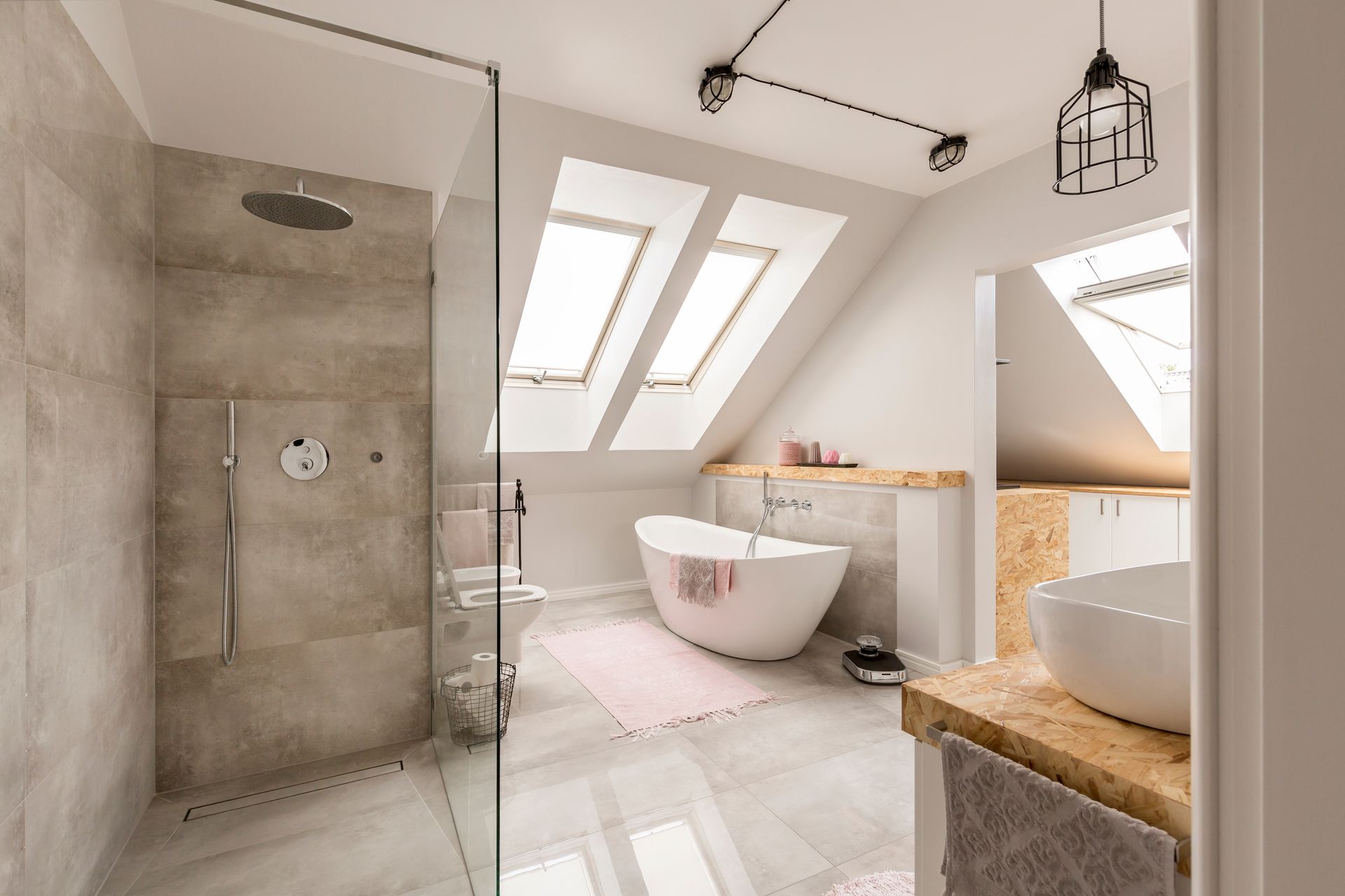 Salle de bains moderne aménagée dans les combles, avec douche à l'italienne, baignoire blanche sur pieds et meuble sous lavabo, sous un puits de lumière.