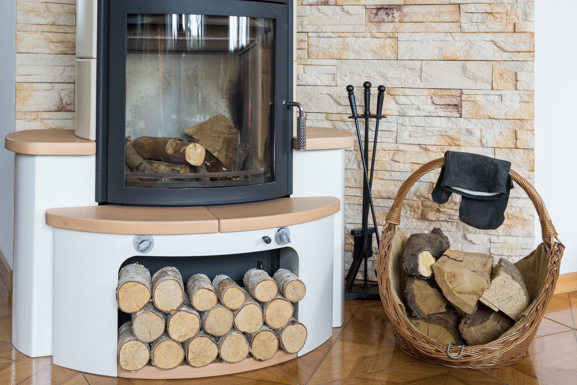 Une cheminée moderne avec un manteau en bois clair, un socle blanc pour ranger les bûches, un ensemble d'outils noirs et un panier en osier rempli de bois.