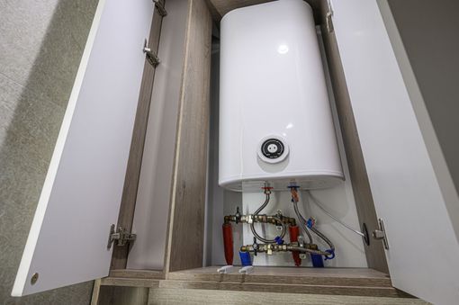Un chauffe-eau électrique blanc installé à l'intérieur d'un meuble mural en bois, laissant apparaître les robinets et les tuyaux de plomberie en dessous.