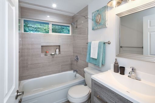 Une petite salle de bains moderne avec une baignoire, un mur carrelé aux tons neutres, des toilettes blanches et un meuble sous lavabo avec miroir.