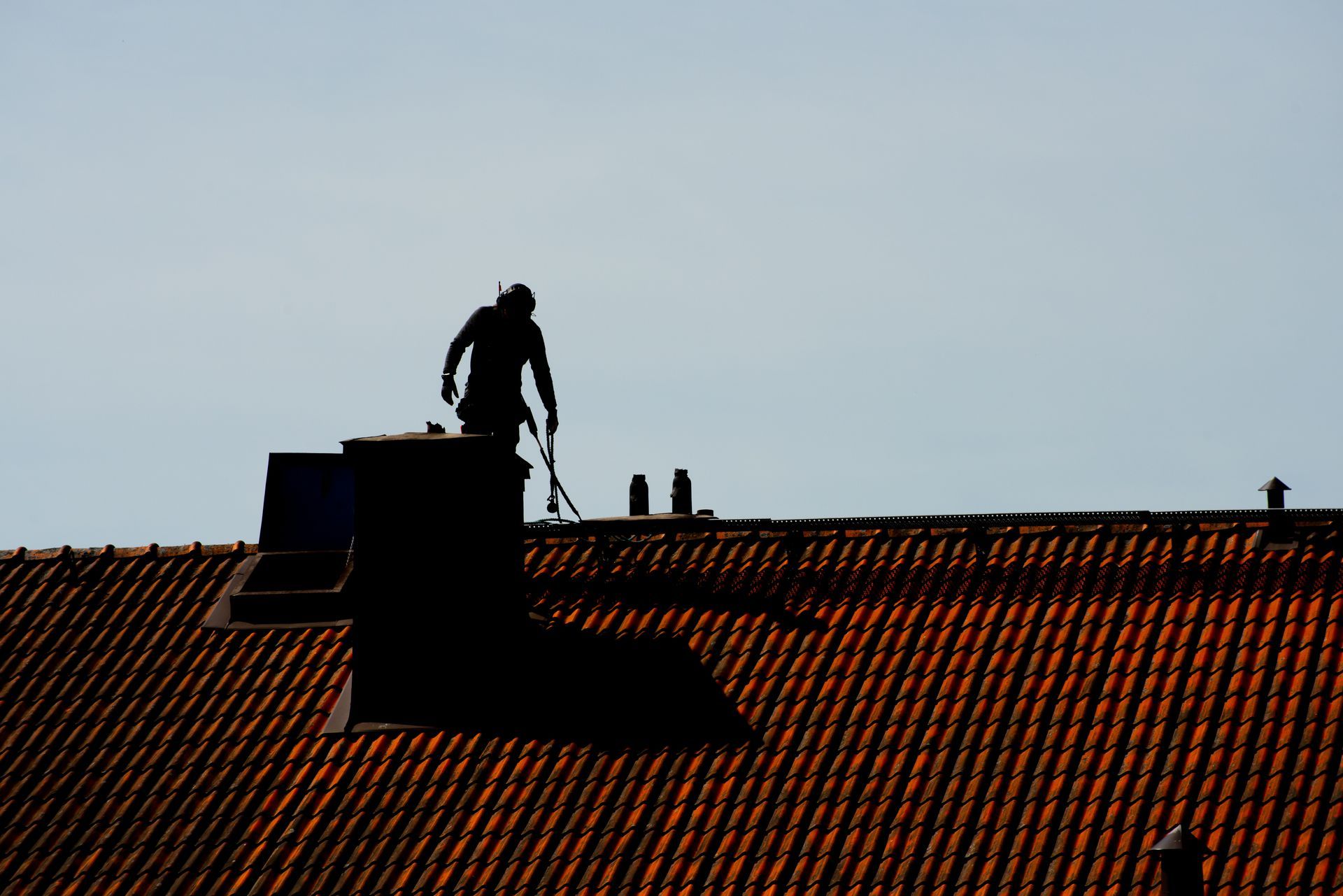 Silhouette d'une personne travaillant sur un toit de tuiles près d'une cheminée, sur fond de ciel clair.