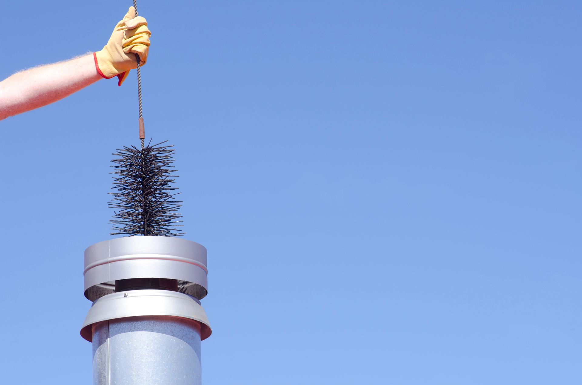Sur fond de ciel bleu clair, une main gantée retire une brosse de cheminée noire d'un conduit métallique.
