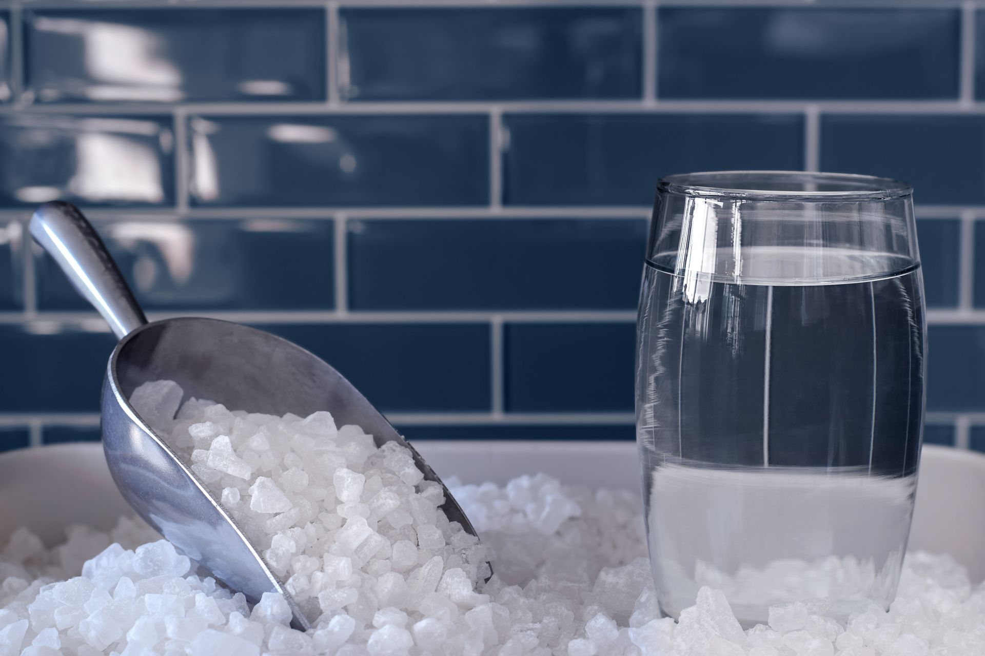 Une pelle en métal remplie de glaçons blancs repose dans un bac à glace à côté d'un verre rempli d'eau, contre un mur carrelé de bleu.