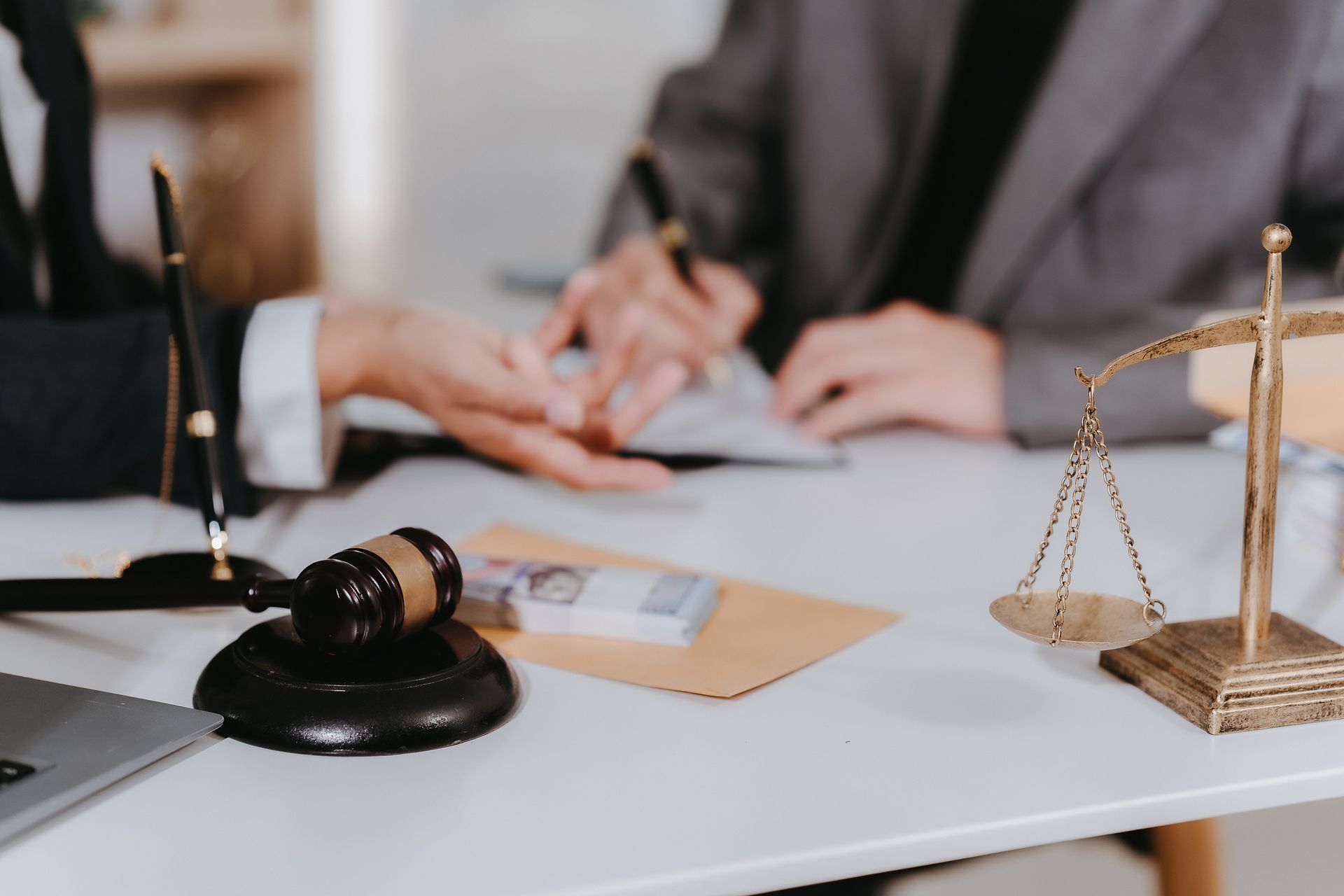 Primer plano de un escritorio con un mazo, una balanza de la justicia y una mano firmando un documento con otra mano ofreciendo.