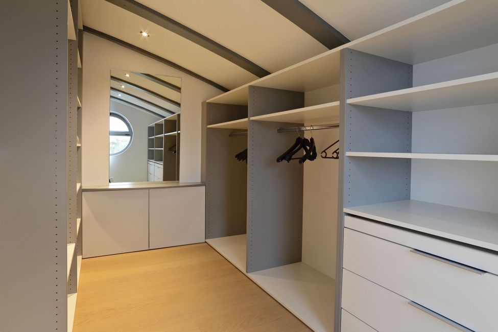 Dressing avec étagères blanches et grises, penderies, tiroirs et plafond voûté. Miroir et fenêtre ronde visibles.