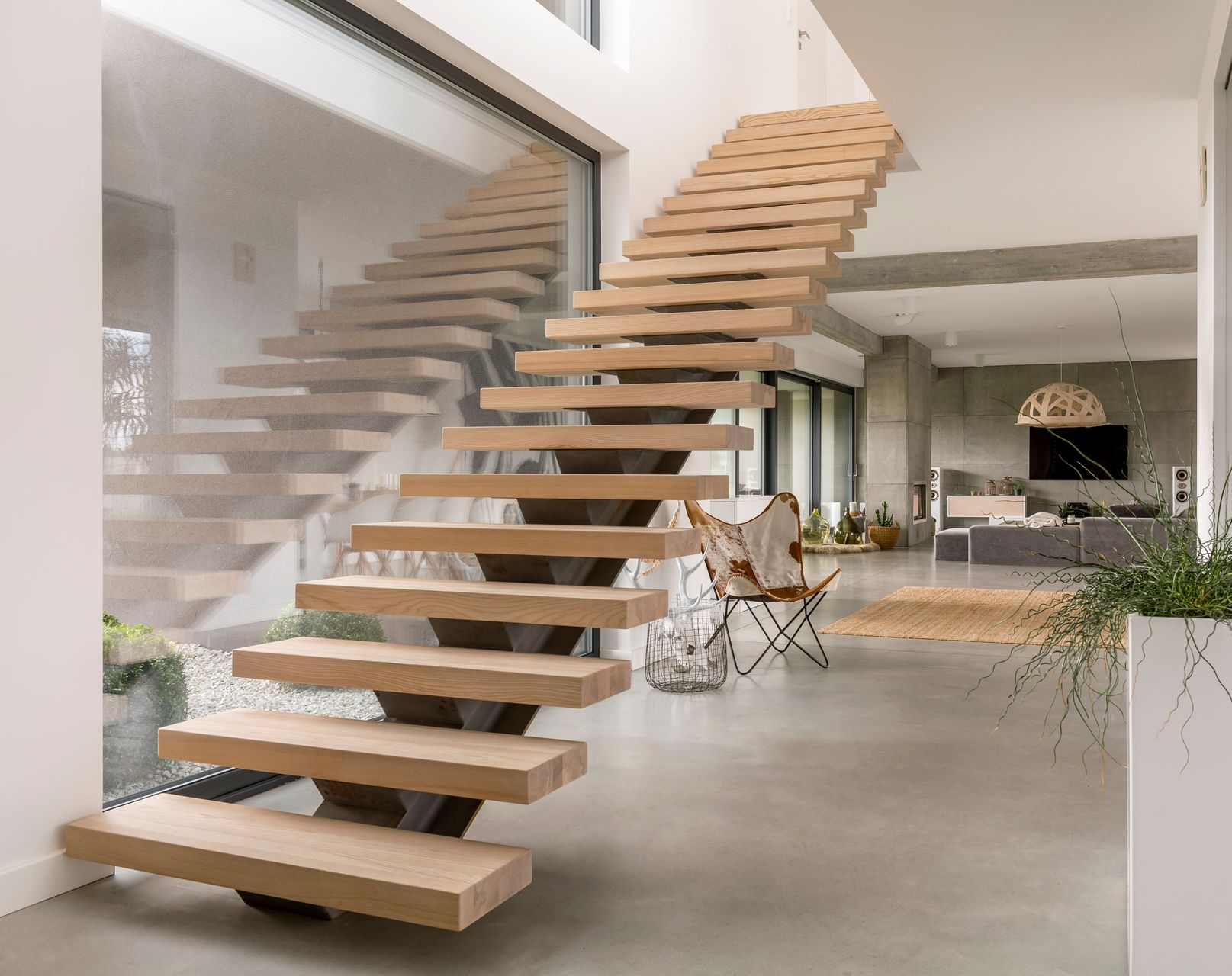 Escalier flottant moderne en bois à l'intérieur d'une maison, avec de grandes fenêtres, des sols en béton et une vue sur le séjour.