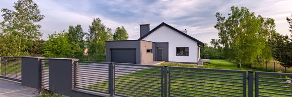 Maison moderne avec garage et toit noir. Une clôture et des arbres se trouvent au premier plan.