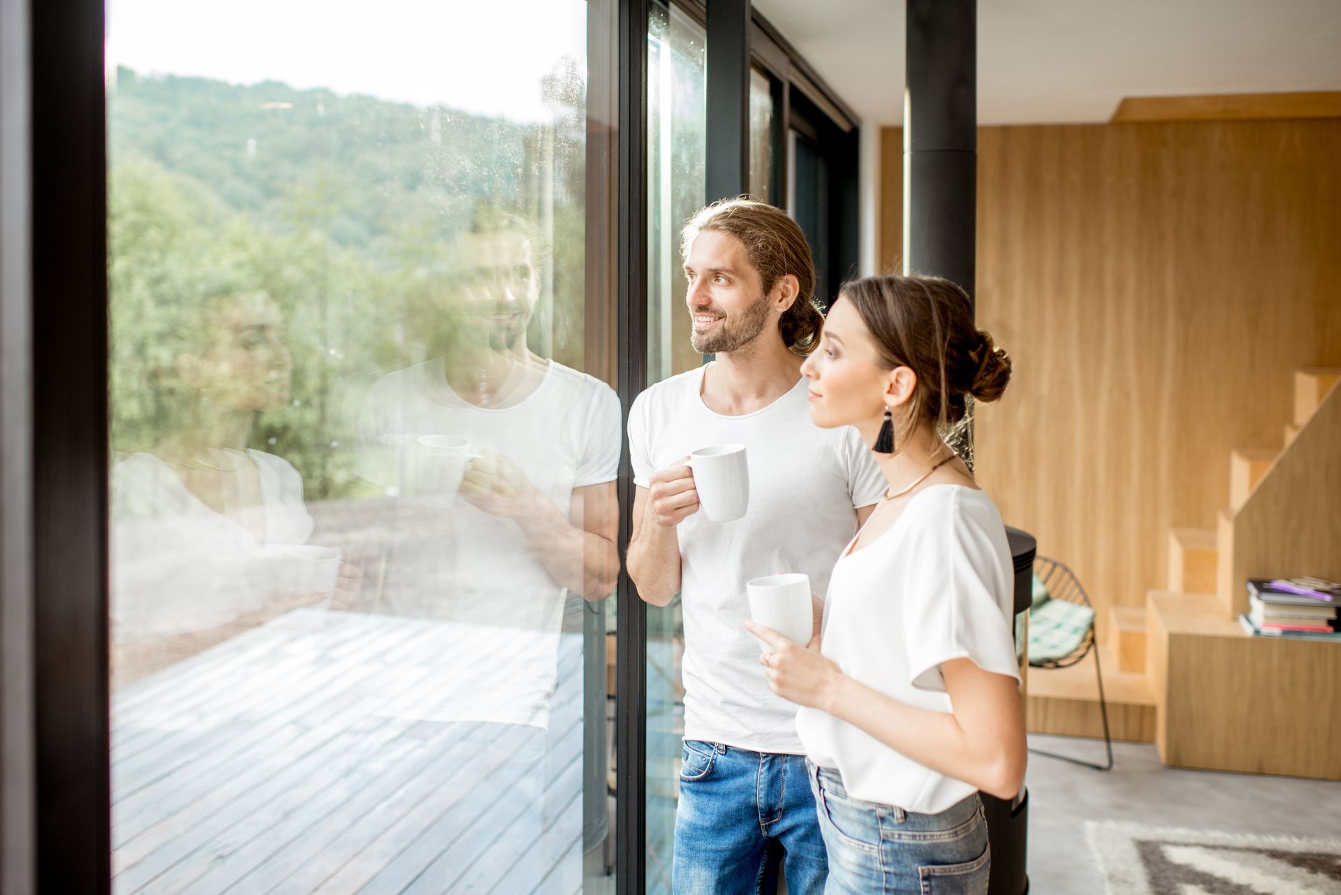 Un couple regardant par la fenêtre, tasses à la main, dans une maison moderne.