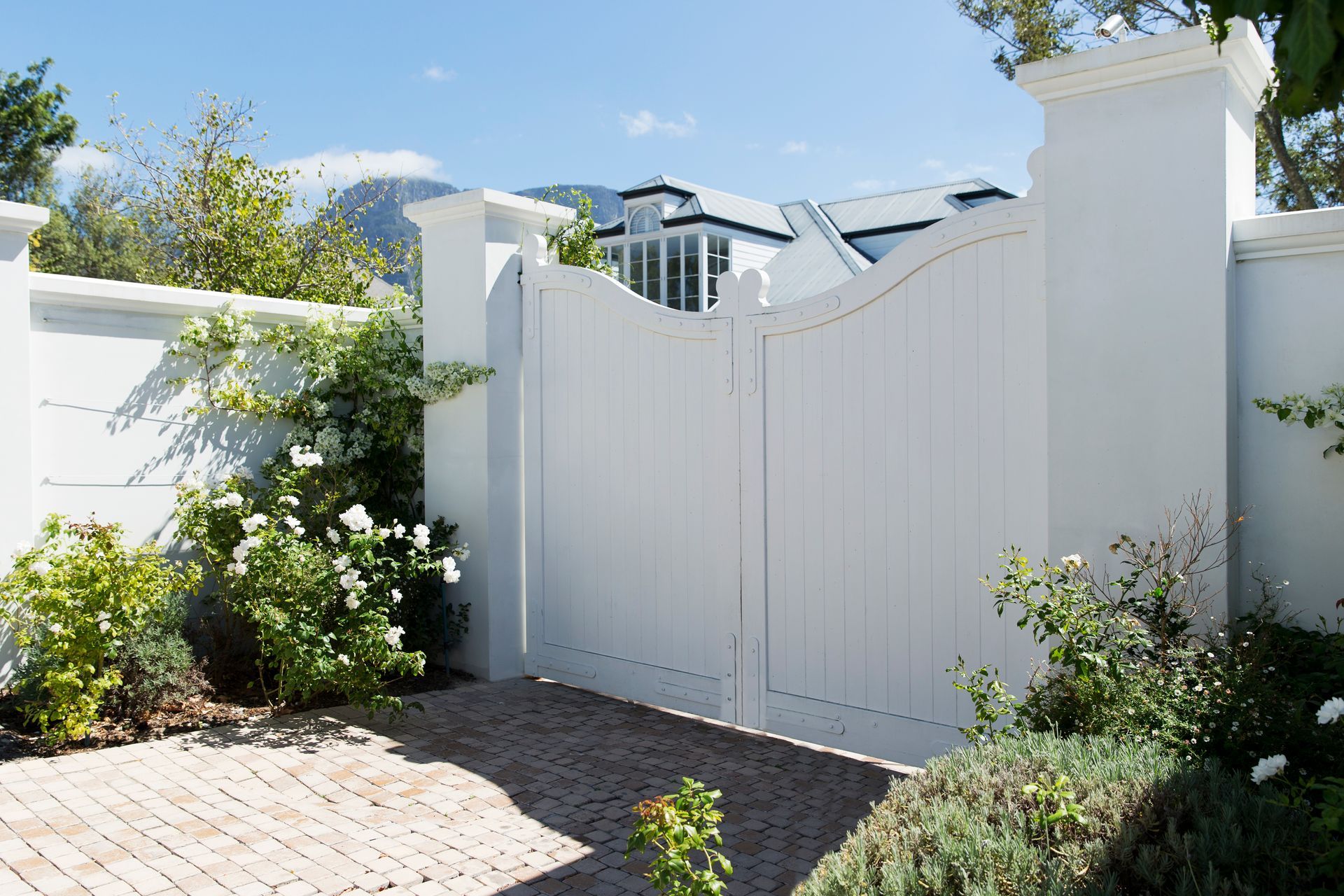 Portail en bois blanc entouré mur blanc et de verdure ; chemin de briques.