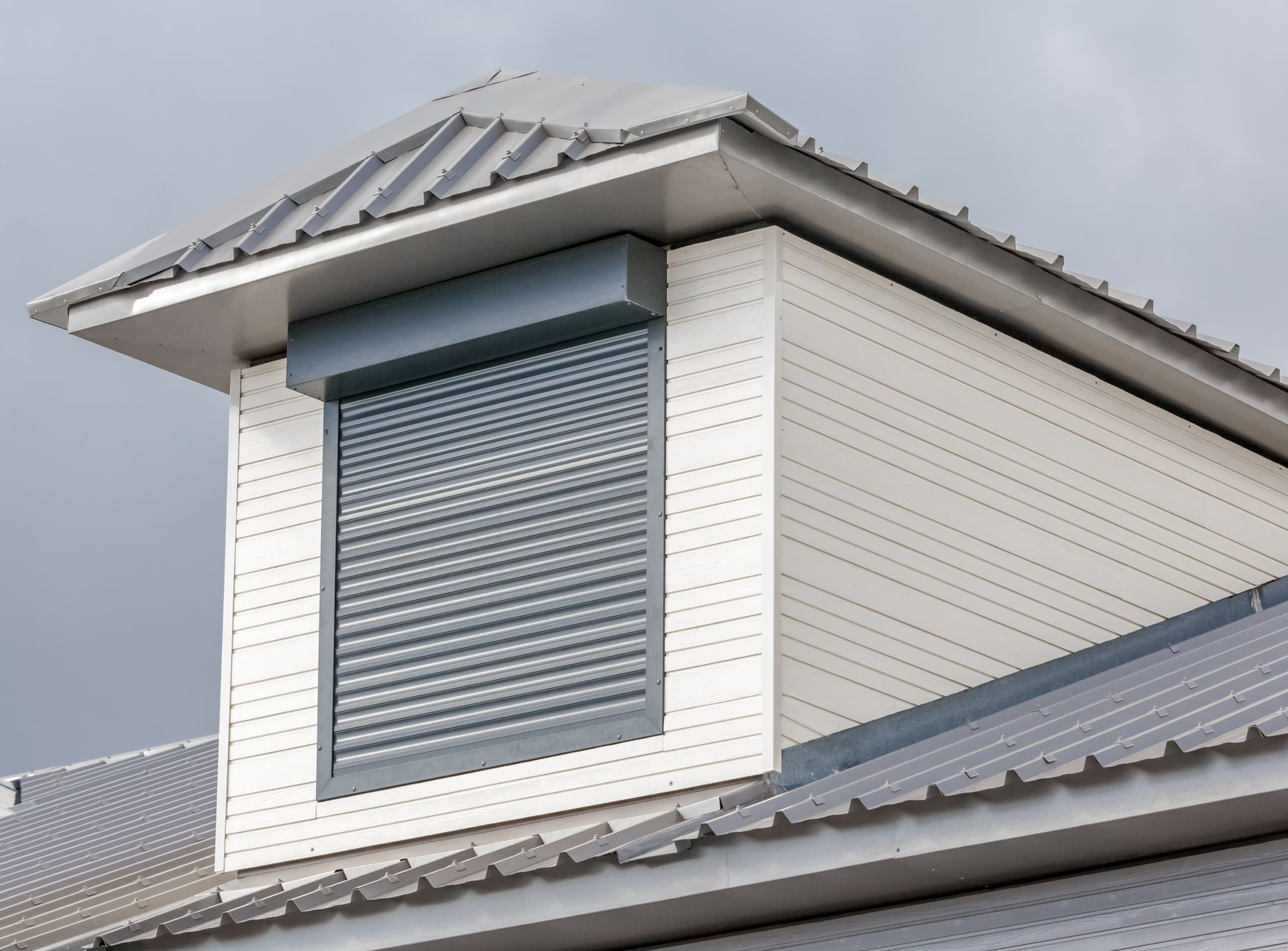 Lucarne avec un volet roulant gris fermé, bardage blanc et toit gris sur fond de ciel nuageux.