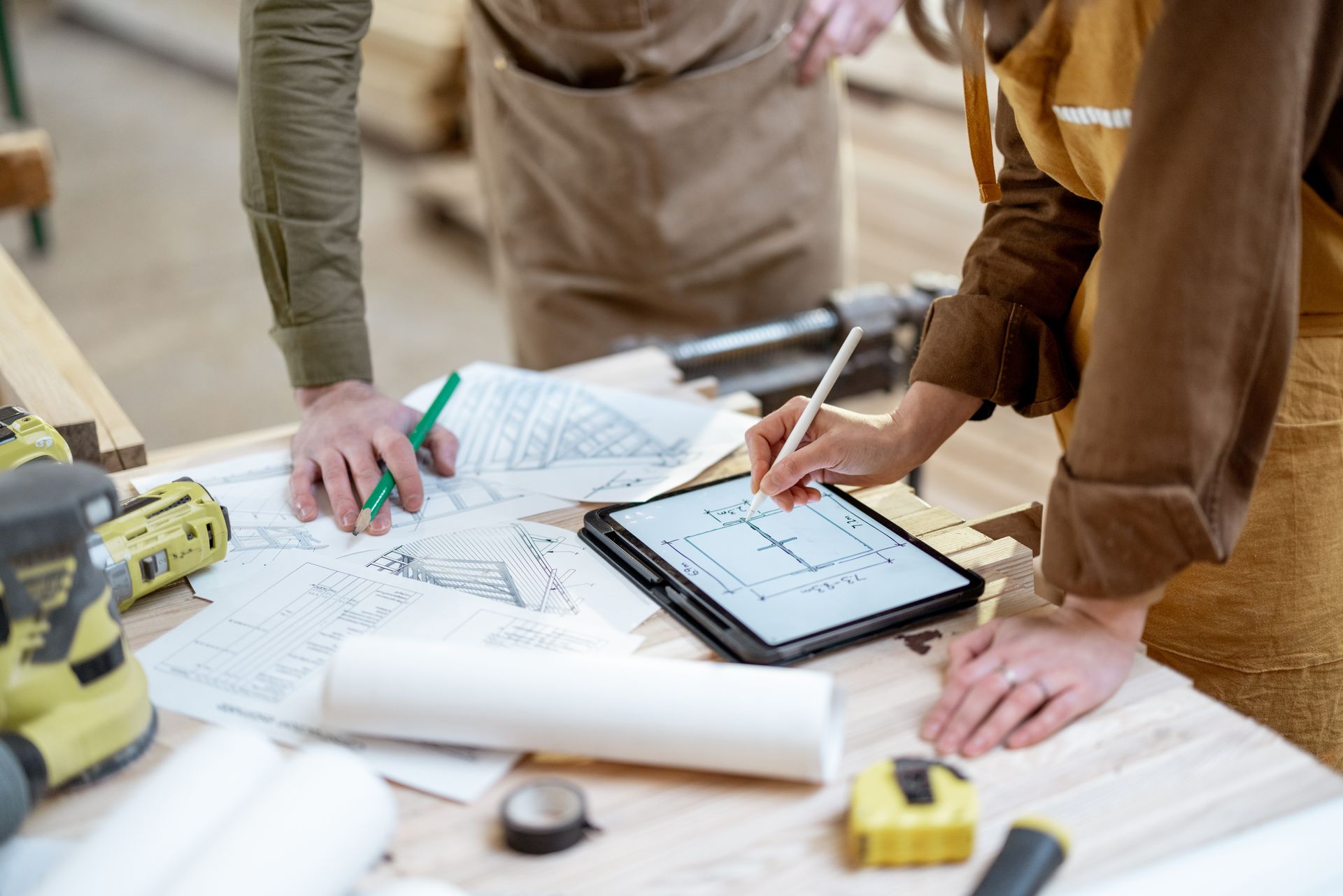 Deux personnes portant des tabliers examinent des plans et une tablette sur un établi dans un atelier.