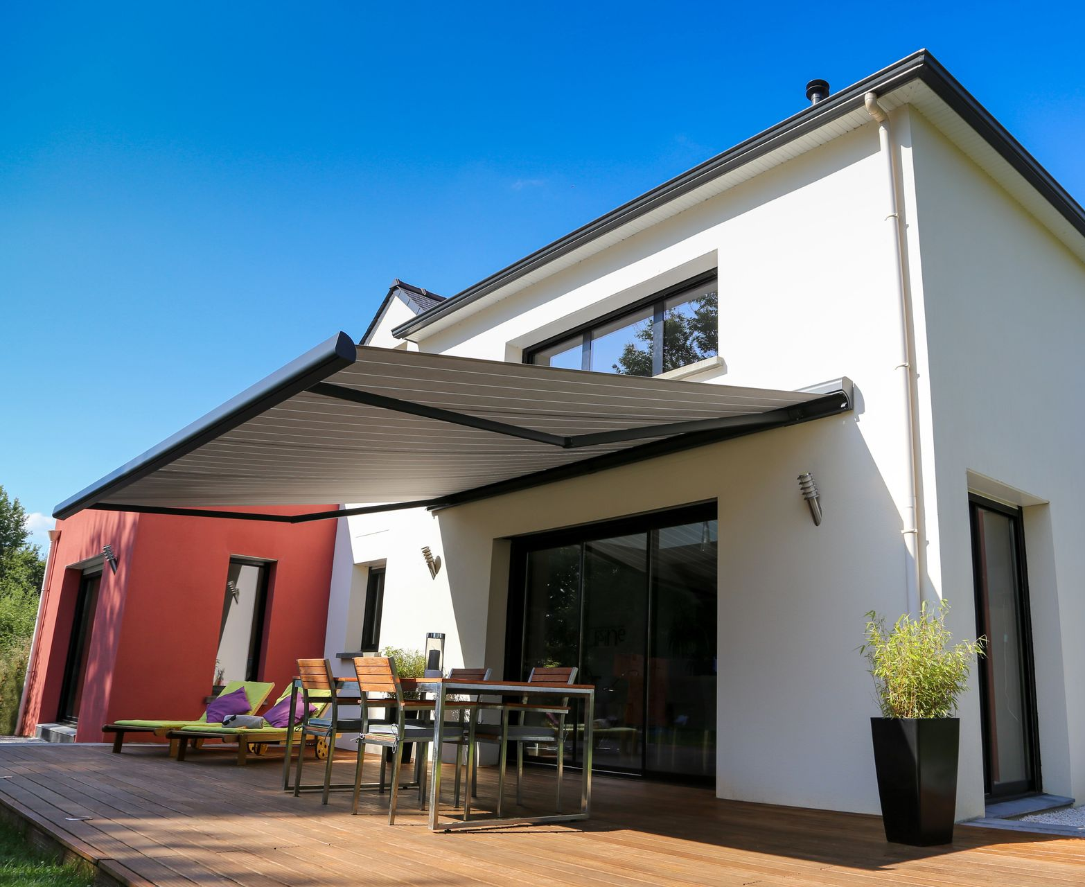Maison moderne avec store rétractable au-dessus du mobilier de jardin sur une terrasse en bois. Journée ensoleillée.