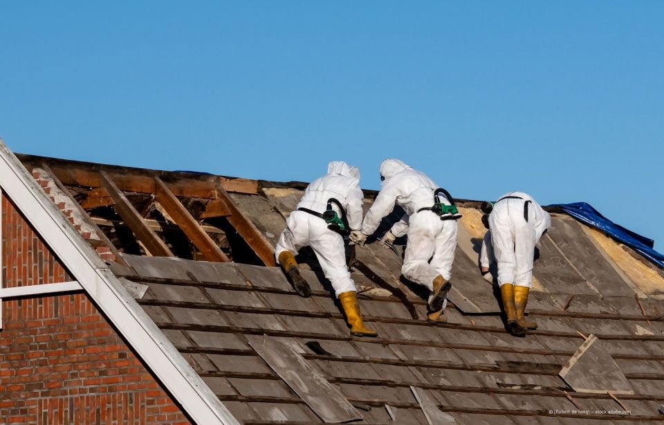Drei Arbeiter in weißen Schutzanzügen reparieren ein beschädigtes Dach mit freiliegenden Dachsparren.