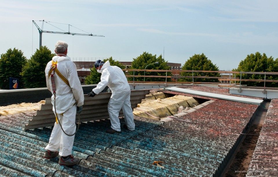 Zwei Arbeiter in weißen Schutzanzügen tragen auf einem Dach eine Beschichtung auf eine blaue Netzoberfläche auf.