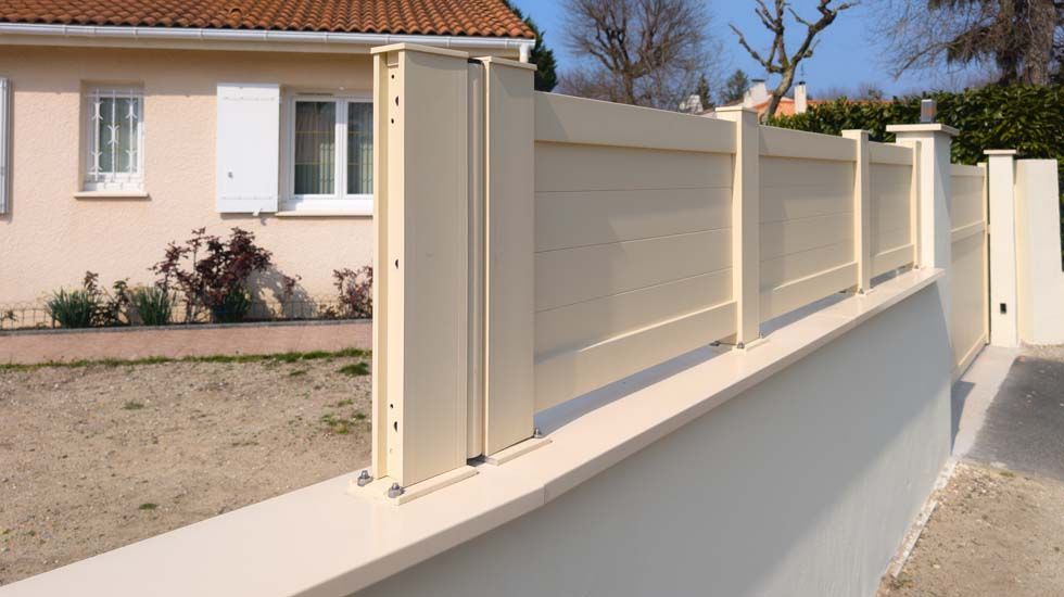 Clôture métallique couleur crème le long d'un mur blanc, à côté d'une maison et d'une allée de gravier.
