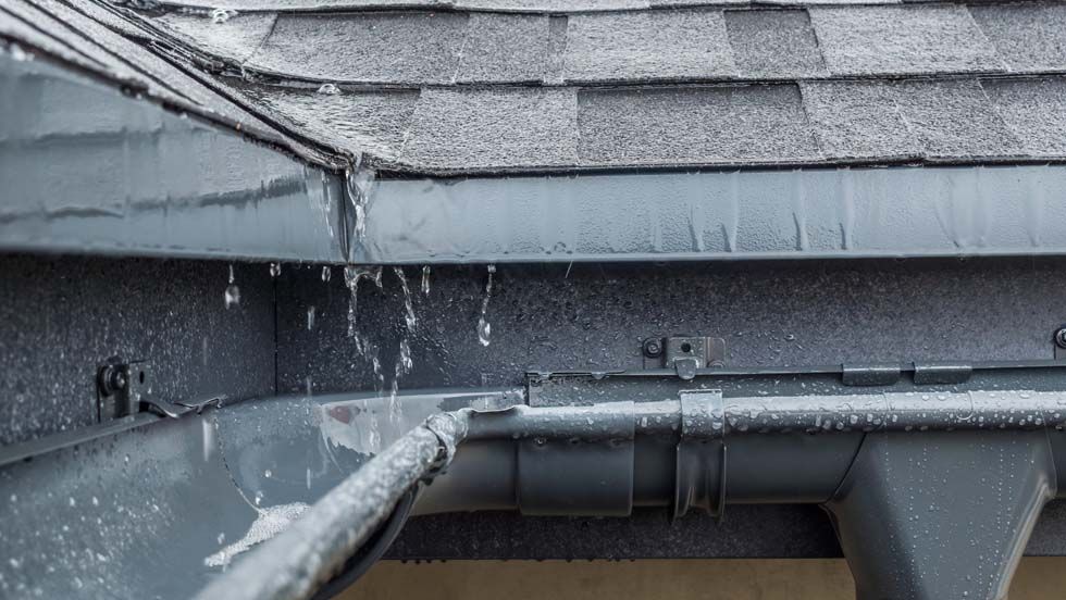 Eau de pluie ruisselant d'une gouttière sur un toit gris et sa descente pluviale.