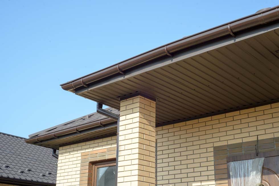 Gros plan sur l'avant-toit d'une maison en briques beiges, avec un soffite brun et un ciel bleu.