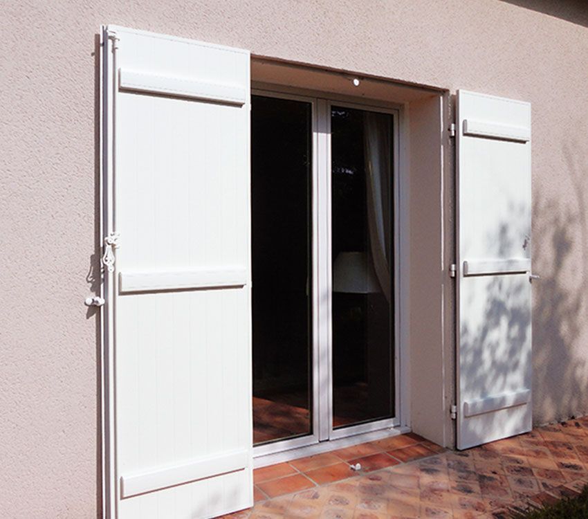 Des volets blancs ouverts encadrent une porte coulissante en verre sur un mur de patio en stuc.